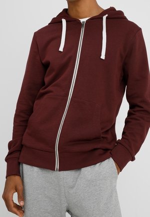 Hombre con sudadera con capucha burdeos y cremallera, con cordones blancos y pantalones deportivos grises, de pie con una mano en el bolsillo.