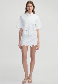 Completo bianco a due pezzi composto da una camicia a maniche corte con bottoni e ritagli e una mini gonna coordinata, entrambi con motivi floreali testurizzati.