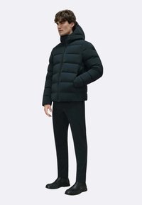 Giubbotto invernale nero con cappuccio, design trapuntato e maniche lunghe. Indossato sopra pantaloni scuri e stivali neri alla caviglia, su uno sfondo neutro.
