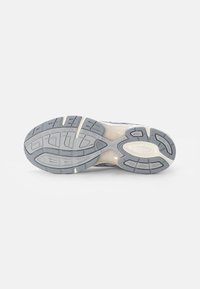 Zapatilla de running con una suela de goma gris texturizada, que presenta una entresuela de color crema, surcos con patrón y una marca acentuada en el talón.