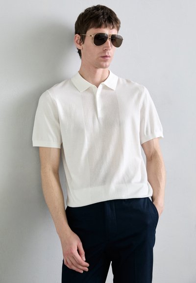 Vitt pikétröja med korta ärmar, strukturerat mönster, rak krage och rullad fåll. Kombineras med mörka byxor och solglasögon.