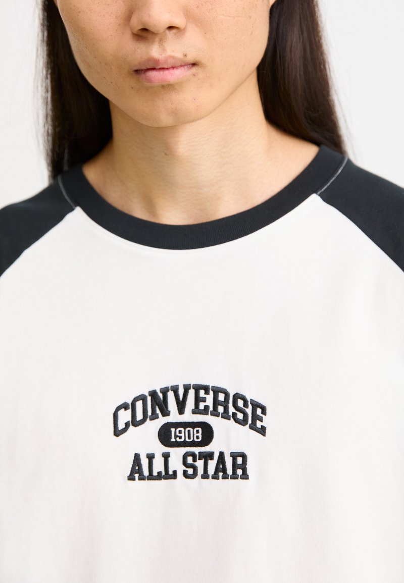T-shirt en coton blanc avec des manches raglan noires, arborant "CONVERSE 1908 ALL STAR" brodé en noir sur la poitrine.