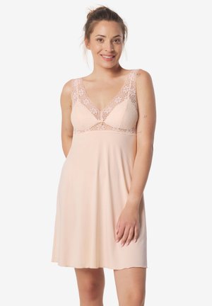 Uśmiechnięta kobieta z włosami związanymi w kok, ubrana w bezrękawnikową, jasnoróżową koronkową koszulkę nocną, stojąca z jedną ręką spoczywającą blisko uda.