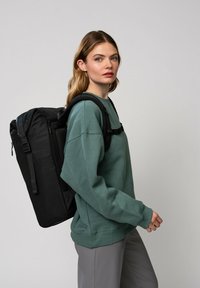 Czarny plecak z teksturowanej tkaniny, z wyściełanymi paskami i kieszenią boczną, noszony na zielonej bluzie. Model stoi na jasnym tle.