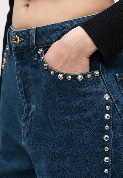 Χέρι στην τσέπη σκούρων μπλε τζιν με χρυσά και διάφανα διακοσμητικά πετράδια κατά μήκος των άκρων της τσέπης και της πλευρικής ραφής, φορεμένα με μαύρο ριμπ μανίκι.