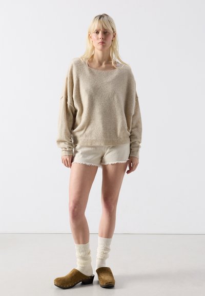 Jeune femme debout portant un pull ample beige, des shorts blancs effilochés, des chaussettes crème et des sabots en daim marron sur un fond blanc uni.