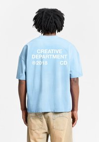 Light blue oversized t-shirt featuring white text: "CREATIVE DEPARTMENT ®2018 CD" on the back. Short sleeves and a relaxed fit.