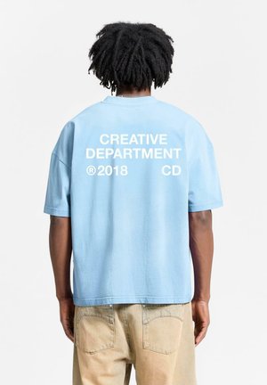 Light blue oversized t-shirt featuring white text: "CREATIVE DEPARTMENT ®2018 CD" on the back. Short sleeves and a relaxed fit.