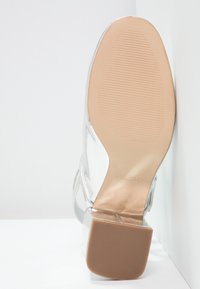 Sandalo con tacco trasparente caratterizzato da una parte superiore trasparente, eleganti accenti argento, punta rotonda e suola beige texturizzata.