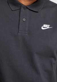 Czarna koszulka polo Nike z dwoma czarnymi guzikami i haftowanym białym logo Nike po lewej stronie klatki piersiowej.