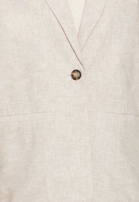 Svetle béžový lněný blazer s jedním kulatým knoflíkem, dvěma předními kapsami a texturovanou tkaninou. Přiléhavý střih s klopami.