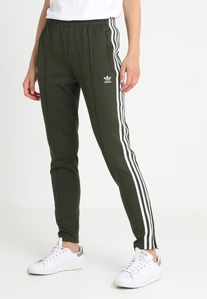 Persona con pantalones deportivos Adidas de color verde oliva con rayas blancas y zapatillas blancas, de pie sobre un fondo blanco.