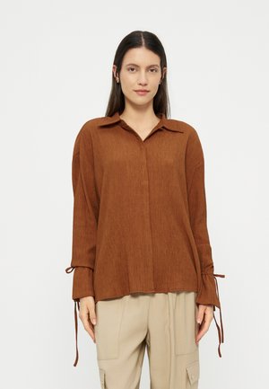 Frau trägt ein lockeres braunes Langarmhemd mit gebundenen Manschetten und beige Cargo-Hosen, steht vor einem schlichten hellen Hintergrund.