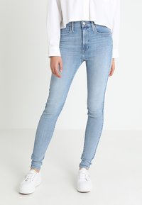 Persona con jeans ajustados de color azul claro, camisa de manga larga blanca y zapatillas blancas, de pie contra un fondo blanco liso.