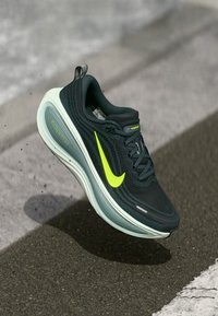 Črne in sive tekaške čevlje z svetlo rumenim Nike swooshom in belim "ZoomX" srednjim podplatom, ki lebdi nad asfaltom in belim cestiškem označevalcem.
