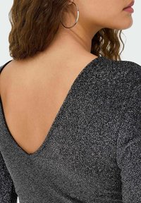 Juodas blizgus palaidinė su gilia V formos nugara, ilgomis rankovėmis ir minkšta tekstūra, šiek tiek prigludusiu siluetu. Matomas sidabrinis lankelio auskaras.