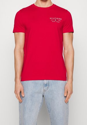 Mężczyzna ubrany w czerwony t-shirt z krótkim rękawem z napisem „Hilfiger Est. 1985 New York” na piersi, w połączeniu z jasnoniebieskimi dżinsami.