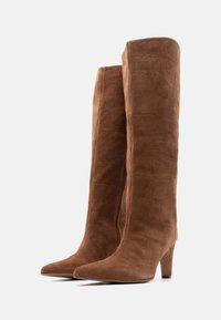 Ζευγάρι ψηλές καφέ μπότες σουέντ με υφή τύπου κροκόδειλου, μυτερές μύτες και μέση ύψους τακούνια σε λευκό φόντο.