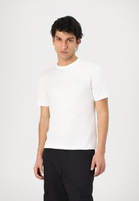 T-shirt bianca in cotone con collo rotondo e maniche corte, caratterizzata da un logo grafico discreto. Indossata con pantaloni cargo neri.