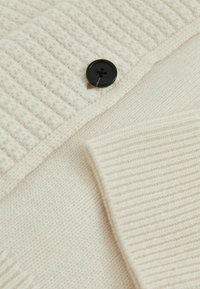 Pull en tricot beige avec un motif en côtes texturé, un tissu lisse et un grand bouton noir comme détail remarquable.