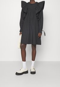 Kvinde iført en sort knælængde kjole med store flæsekantede skuldre, hvide sokker, cremefarvede loafers og holding en lille hvid håndtaske.