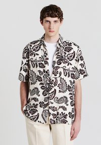 Kurzarmhemd aus cremefarbenem Stoff mit schwarzem Paisleymuster, mit lockerer Passform und Button-Down-Kragen, kombiniert mit beigen Hosen.