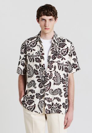 Chemise à manches courtes en tissu crème avec un motif paisley noir, dotée d'une coupe décontractée et d'un col boutonné, assortie à un pantalon beige.