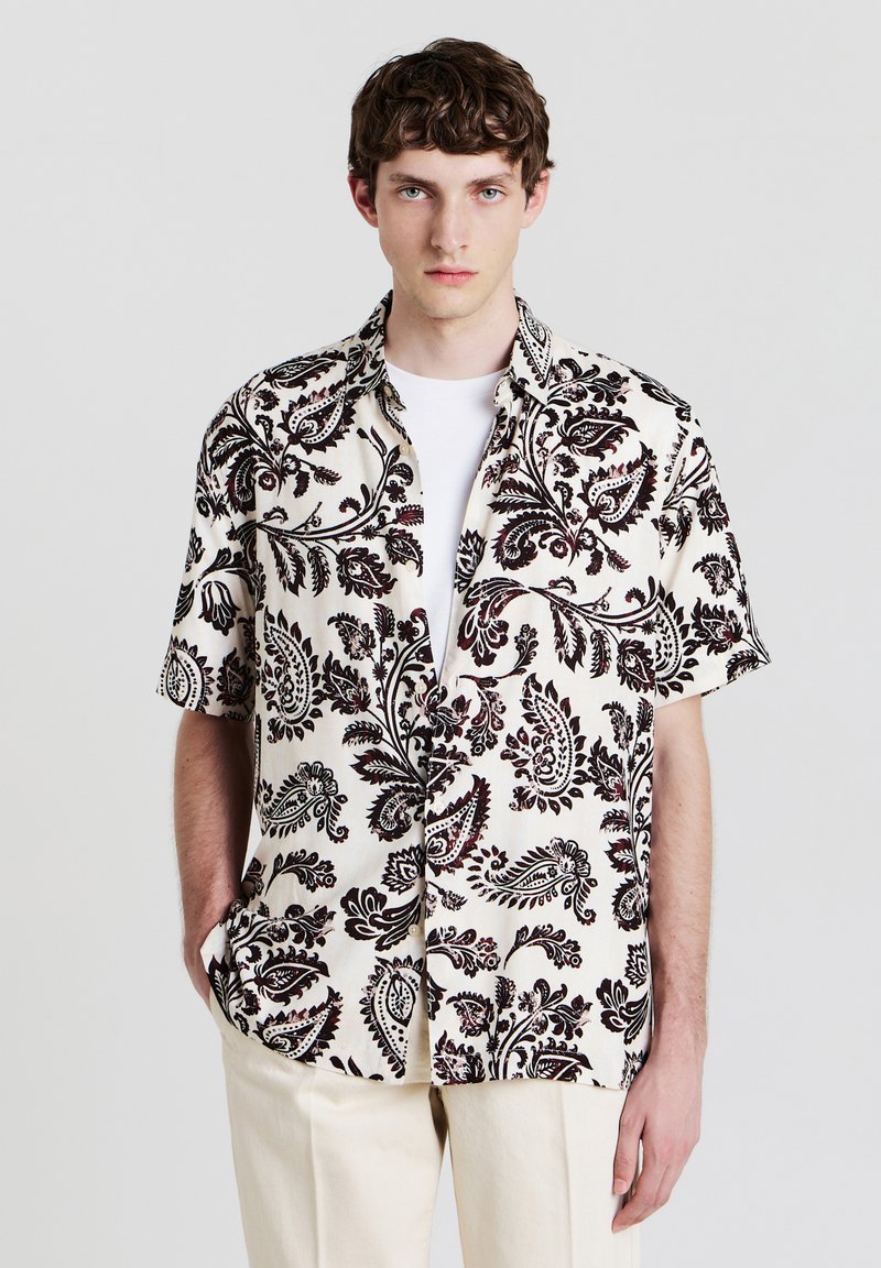 Kurzarmhemd aus cremefarbenem Stoff mit schwarzem Paisleymuster, mit lockerer Passform und Button-Down-Kragen, kombiniert mit beigen Hosen.