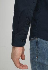Dunkelblauses Hemd mit langen Ärmeln, das zwei schwarze Knöpfe an den Manschetten zeigt; glattes Baumwollgewebe; taillierter Schnitt; getragen mit hellblauen Jeans.