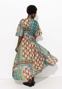 Femme aux cheveux courts et bouclés portant une robe fluide en patchwork aux motifs avec des tons de turquoise, marron et vert, se tenant devant un fond blanc.