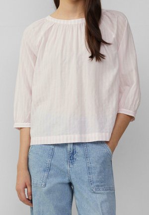 Kvinde iført en lyserød, løstsiddende bluse med knap til under armen og lyseblå højtaljede jeans med store forlommer.