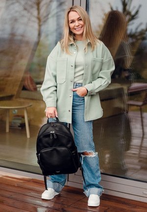Schwarzer Rucksack mit runder Form und doppeltem Reißverschluss, getragen mit einer hellgrünen oversized Jacke, einem hellen Oberteil und zerrissenen Jeans.