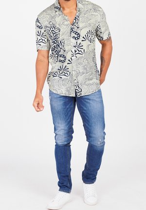 Mężczyzna w krótkim rękawie koszuli zapinanej na guziki z abstrakcyjnym czarno-białym wzorem, niebieskich dżinsach i białych trampkach stoi na tle białego tła.