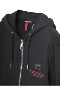 Crna zip-up dukserica s kapuljačom, metalnim zatvaračem, malim četvrtastim logom, i crvenim tekstom na prsima ispod loga.