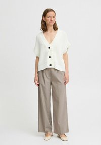 Cardigan bianco lavorato a maglia con scollatura a V profonda, maniche corte e bottoni neri. Abbinato a pantaloni beige a quadri a gamba larga e scarpe chiare.
