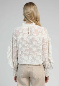 Blouse brodée de fleurs blanches avec un tissu transparent, des manches à volants et une fermeture en forme de clé au dos. Associée à un pantalon beige clair.