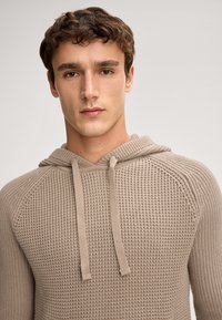 Sweatshirt à capuche beige présentant un motif tricoté texturé, des cordons de serrage réglables et une coupe décontractée avec des manches raglan.