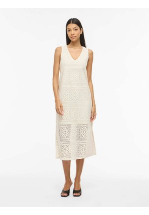 Femme debout portant une robe midi en crochet sans manches de couleur crème avec des motifs floraux et des talons ouverts noirs sur fond blanc.