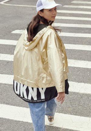 Chaqueta de lluvia metálica dorada con capucha, sección inferior negra con el logotipo "DKNY", que se lleva sobre unos vaqueros azules y calzado ligero.