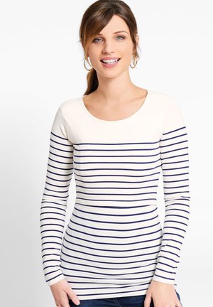 Smiling woman with brown hair in a ponytail wearing hoop earrings and a white long-sleeve shirt with navy horizontal stripes.