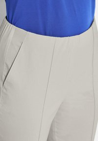 Beige Hosen mit elastischem Bund und seitlichen Taschen, getragen mit einem in die Hose gesteckten blauen Hemd, zeigen die vorderen und seitlichen Nahtdetails.