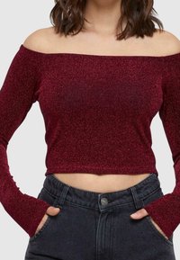 Viininpunainen off-the-shoulder cropped-toppia pitkähihaisella, joka on valmistettu kiiltelevästä kankaasta. Yhdistetty tummiin korkea vyötäröisiin farkkuihin, joissa on etutaskut.