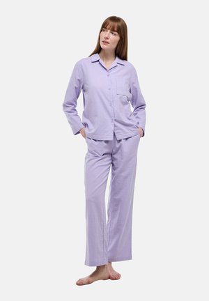 Femme debout pieds nus portant une chemise boutonnée à manches longues violet clair et un pantalon large assorti sur fond blanc.