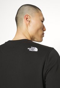 Sudadera deportiva negra con cuello redondo y puños acanalados. Logo blanco notable en el hombro derecho. Textura suave.