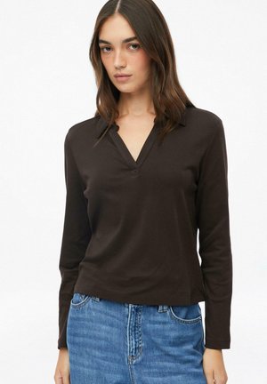 Junge Frau trägt ein dunkelbraunes langärmeliges V-Ausschnitt-Shirt und blaue Jeans und steht vor einem schlichten weißen Hintergrund.