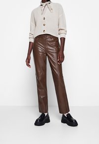 Beige stickad kofta med stora knappar, bärs över en ljus topp. Bruna läderbyxor och kraftiga svarta skor fulländar outfiten.