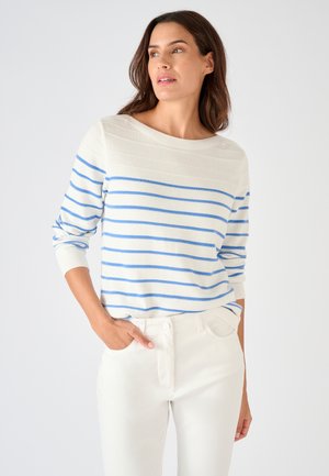 Mujer con suéter blanco con rayas horizontales azul claro y pantalones blancos, de pie con una mano en el bolsillo, mirando ligeramente hacia la derecha.