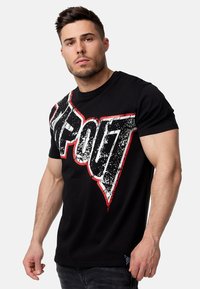 Camiseta de algodón negra con un gran gráfico desgastado de "TAPOUT" en blanco y rojo colocado en diagonal en la parte delantera. Mangas cortas.