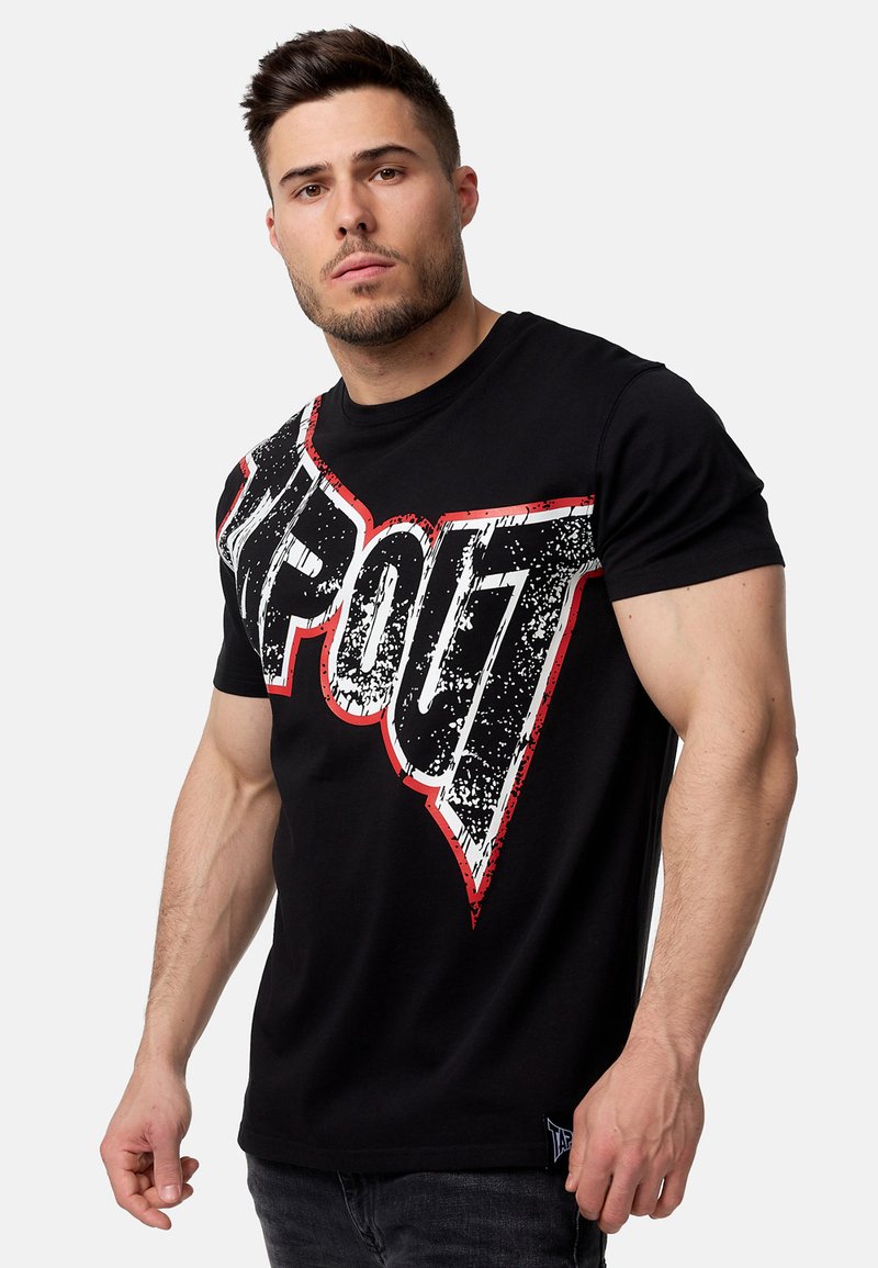 Camiseta de algodón negra con un gran gráfico desgastado de "TAPOUT" en blanco y rojo colocado en diagonal en la parte delantera. Mangas cortas.