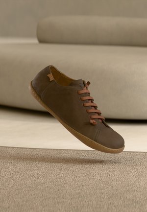 Brauner Lederschuh mit braunen Schnürsenkeln, minimalistischem Design, leicht abgerundeter Spitze und dünner Gummisohle, schwebend über einer strukturierten Oberfläche.